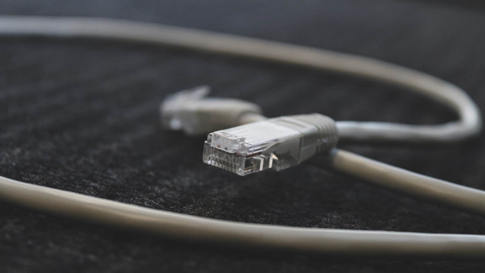 Close-up of an unplugged Ethernet cable with a clear connector lying on a textured black surface. The cable is beige and slightly curved, with the unplugged end in focus and the rest of the cable blurring into the background.