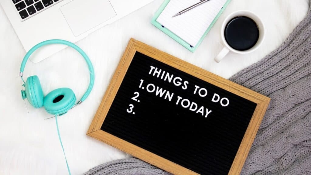 A list of tasks on a blackboard next to a cup of coffee, headphones, and laptop. The text on the blackboard reads: “Things to do: 1. Own today”.