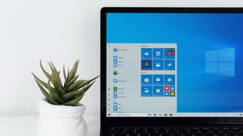 An open laptop on a desk displaying the Windows Start Menu with various application tiles such as Mail, Calendar, Microsoft Edge, and Photos visible. To the left of the laptop is a potted succulent plant on a white surface, adding a touch of greenery to the workspace.