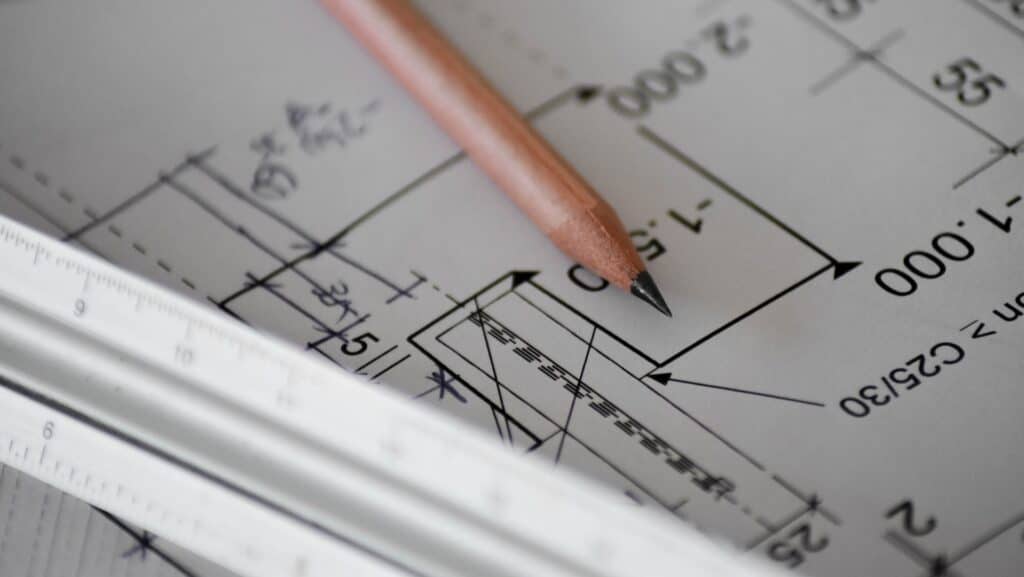 A close-up photo of an architectural blueprint featuring various measurements and design details. A wooden pencil rests on the paper, pointing to a specific section of the plan, with a metal ruler lying parallel to the pencil.