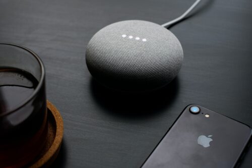 A grey Google Nest Mini smart speaker placed on a table, next to an iPhone and cup of coffee.