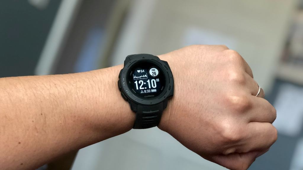 A person's wrist wearing a black Garmin sports watch. The watch face displays the time as 12:10, along with the date 'Fri 26' and a heart rate reading of '64'. There's also a battery indicator and Bluetooth symbol shown at the top of the watch face. The background is blurred, focusing attention on the watch.