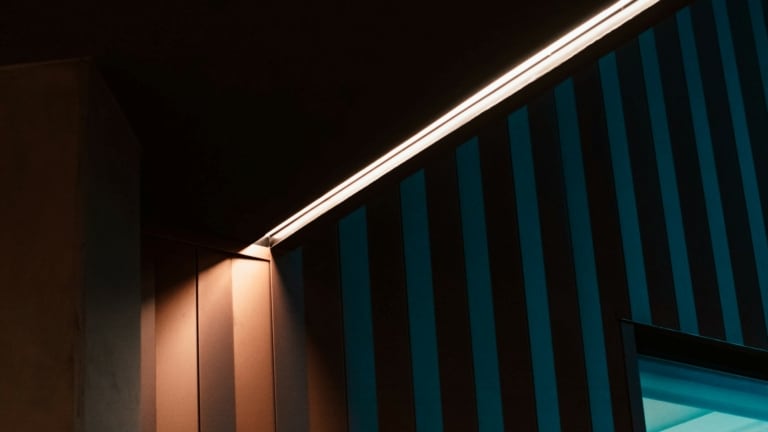 An abstract view of a room corner where a warm LED strip light runs along the edge where the ceiling meets the wall. The wall has vertical teal and black stripes, creating a striking contrast with the warm light. The lower part of the image shows a window emitting a soft blue light, adding a layer of depth and colour contrast to the scene.