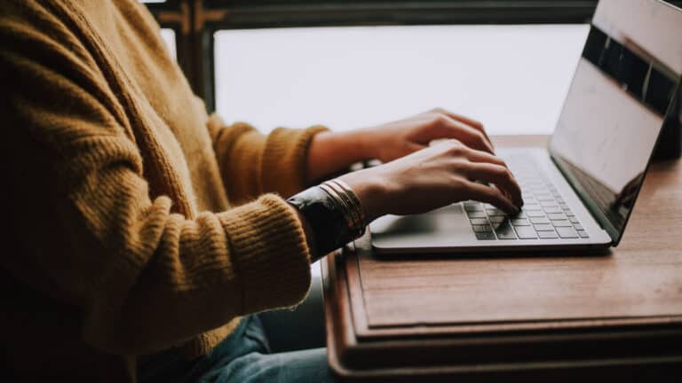 A person in a mustard yellow sweater is typing on a silver laptop placed on a wooden table. The focus is on the person's hands, which are adorned with bracelets, and the laptop keyboard. The background is softly lit, and a window can be seen, suggesting a cozy indoor setting.